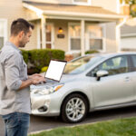 Peut-on estimer sa voiture sans fournir d’adresse électronique ? Un propriétaire consulte un ordinateur portable près d’une voiture compacte stationnée devant une maison lumineuse.