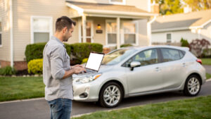 Un propriétaire consulte un ordinateur portable près d’une voiture compacte stationnée devant une maison lumineuse.