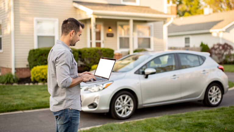 Un propriétaire consulte un ordinateur portable près d’une voiture compacte stationnée devant une maison lumineuse.